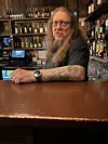 Bartender med langt hår og tatoveringer står bak en brun bardisk foran flasker i hyller.